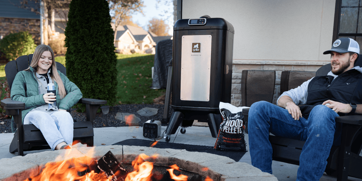 a couple sitting at a firepit with a fire going with the grilla mammoth smoking in the background and wood pellets sitting next to the grilla mammoth vertical pellet smoker